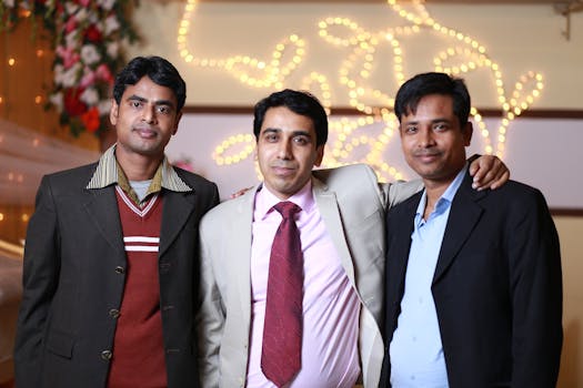 Three men in formal attire smiling together at an indoor event with festive lighting.