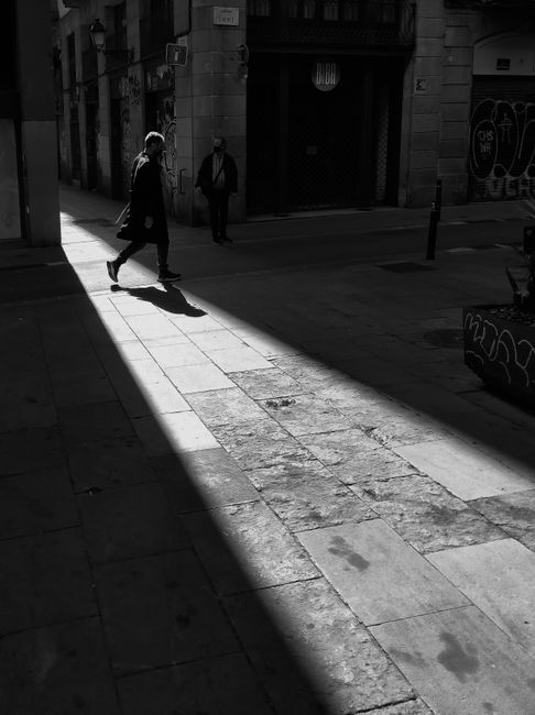 Moody black and white shot of people walking on a Barcelona street with dramatic shadows.