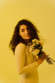 A woman with curly hair in a white dress holds a bouquet against a yellow backdrop.