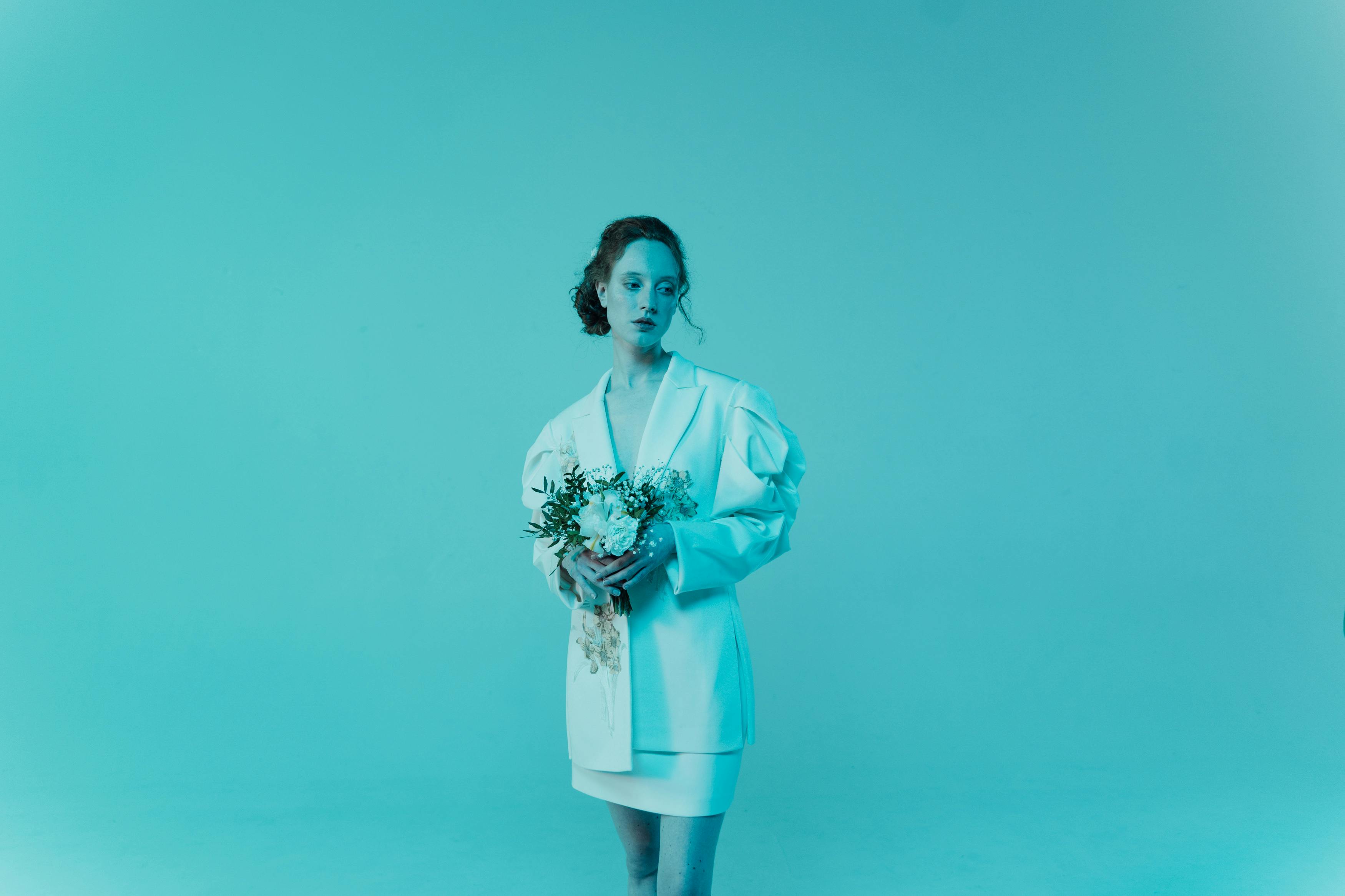 Free An artistic photo of a woman in a white suit holding a floral bouquet, set against a cool-toned backdrop. Stock Photo