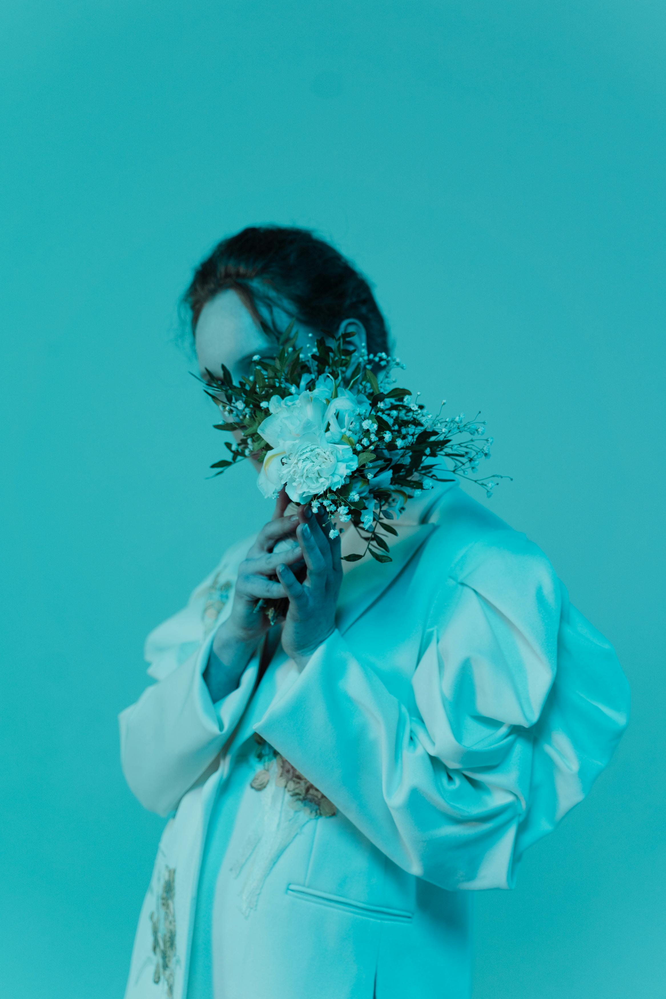 Woman in white holding flowers against a blue backdrop, face obscured for privacy.