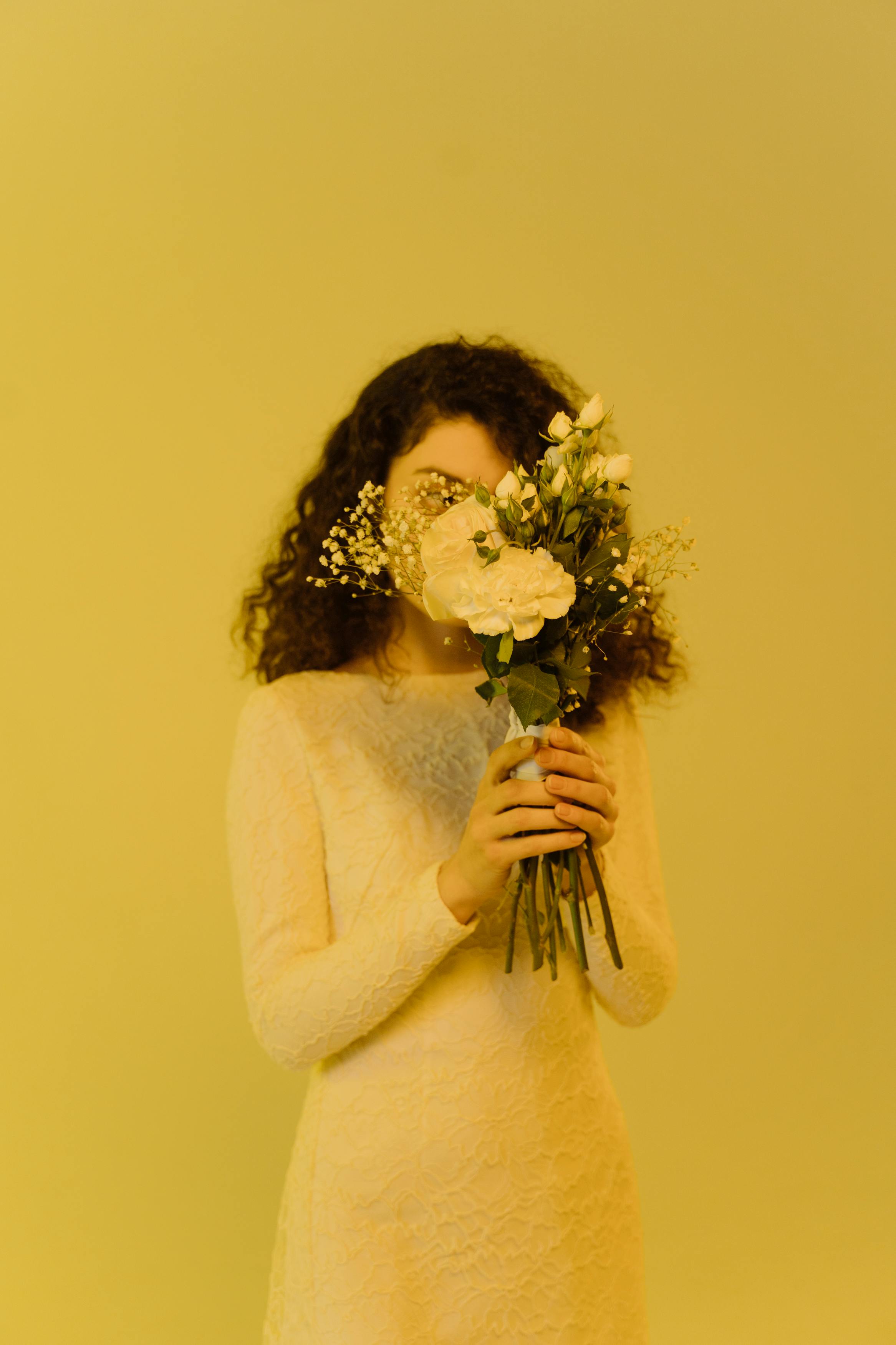Free Woman in white lace dress holding bouquet hiding her face on yellow background. Stock Photo