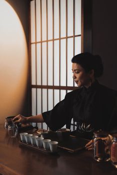 An Asian woman gracefully prepares a traditional tea ceremony indoors. Warm lighting enhances the intimate setting.