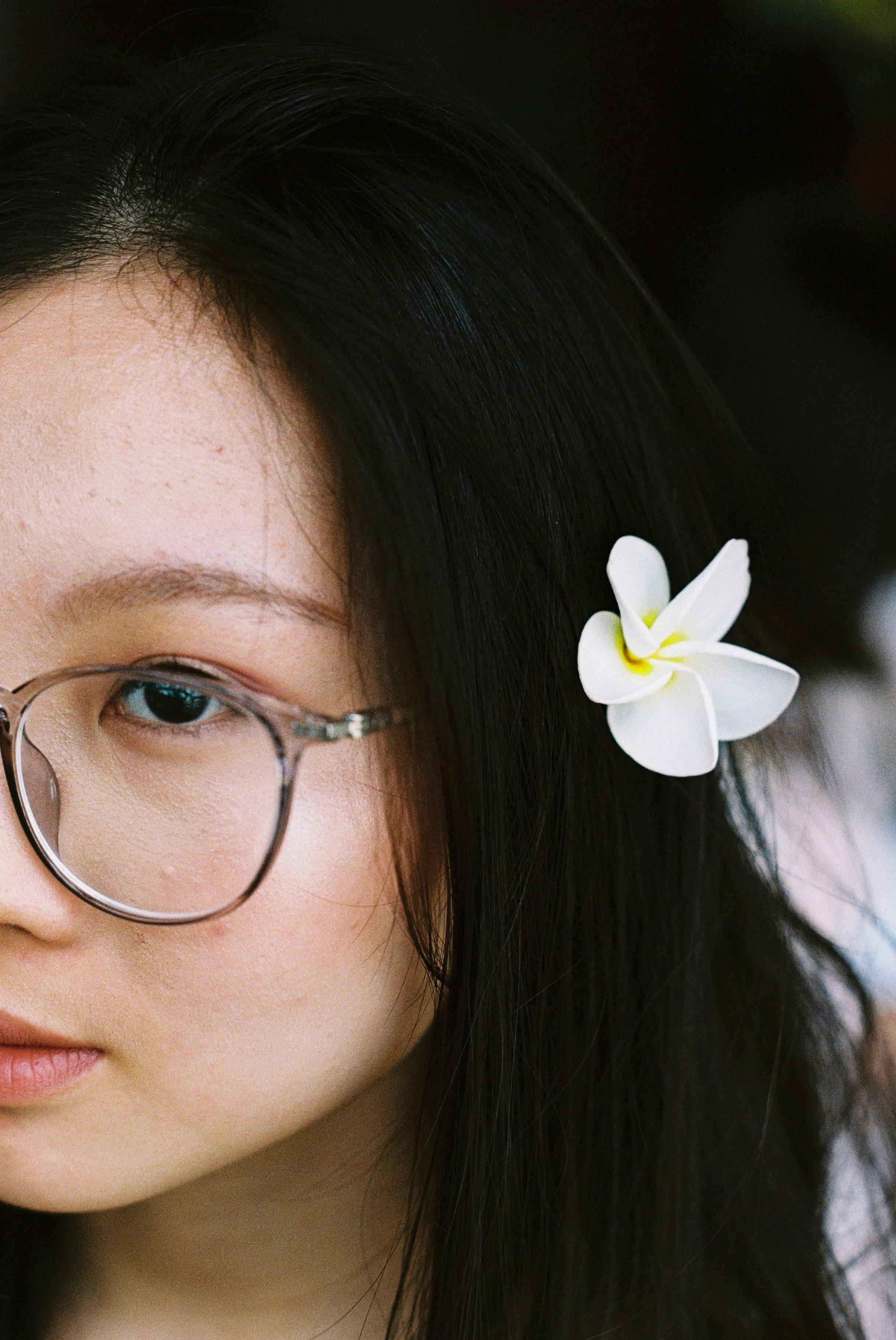 Close-up of a young woman wearing glasses with a white flower in her hair.