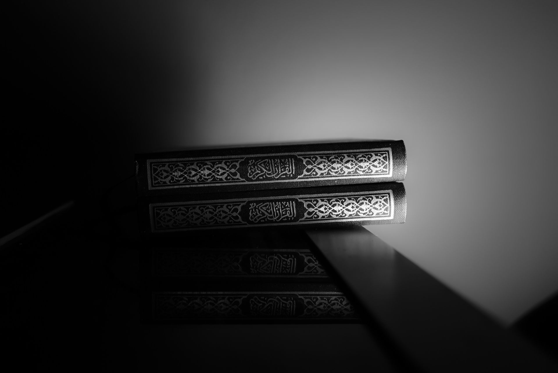 Monochrome image of two Qurans in Arabic script on a reflective surface, symbolizing spirituality and religion.
