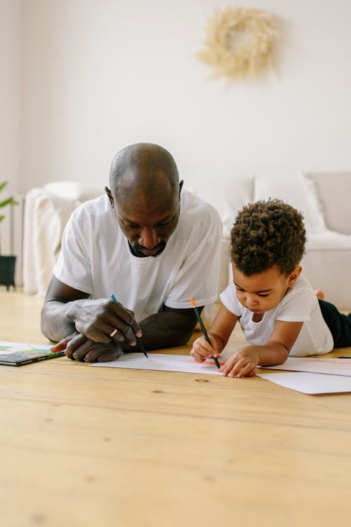 Free A Man Lying Down on the Floor with His Son while Drawing on Paper Stock Photo