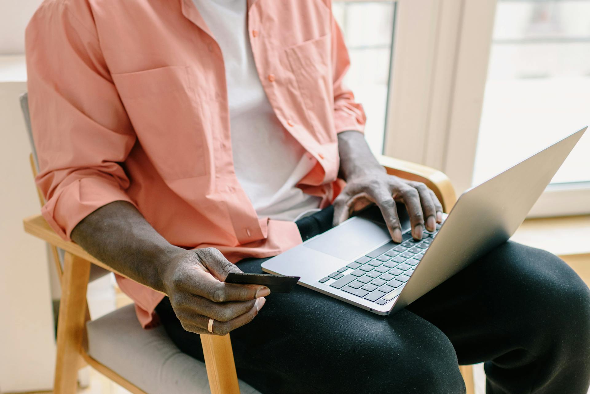 A man sitting with a laptop and credit card, browsing online indoors during the day.