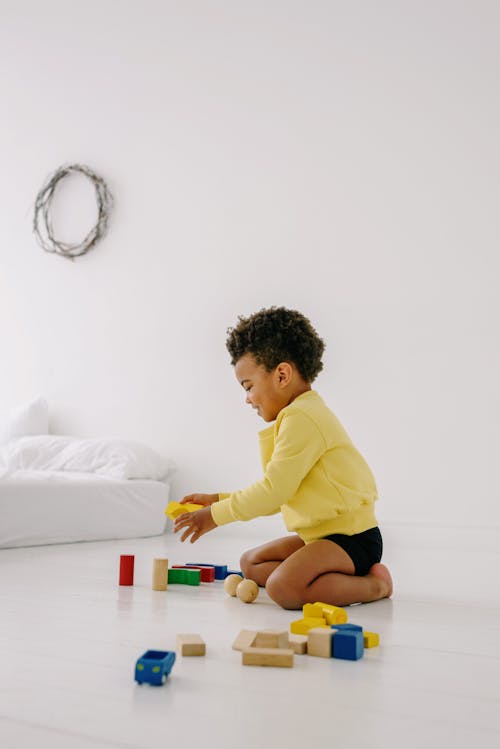 A Boy in Yellow Sweater Sitting on the Floor while Playing Toy Blocks