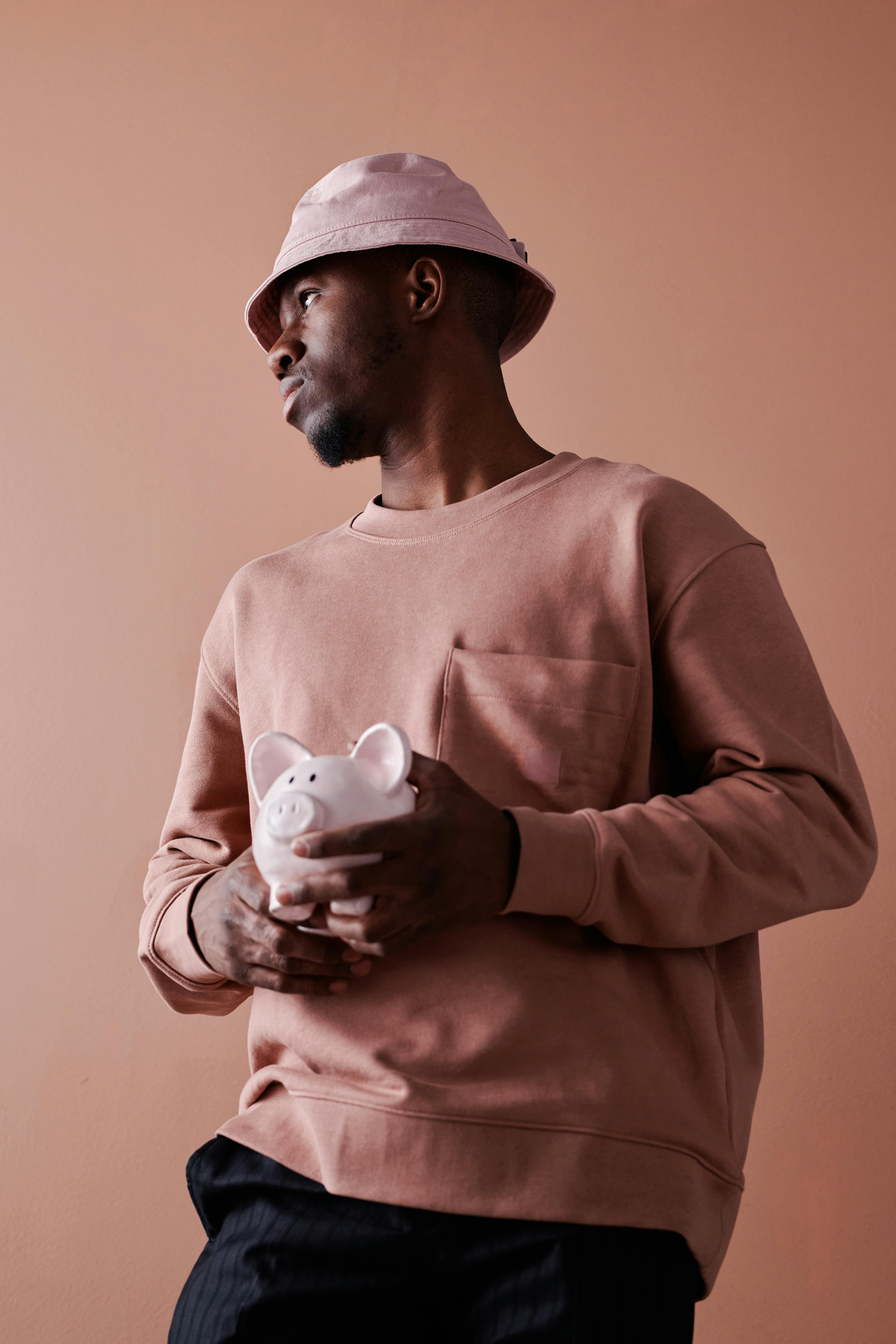 A man in a bucket hat holds a pink piggy bank indoors, embodying savings and financial planning concepts.