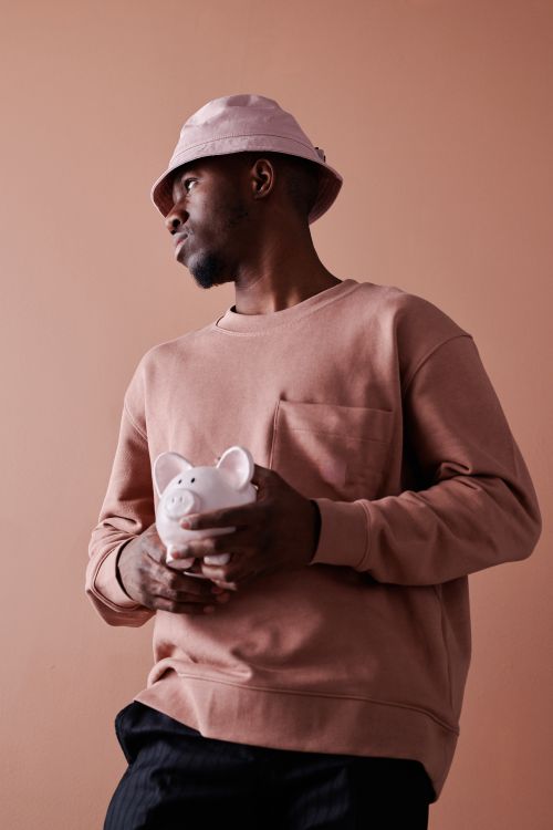 Free A man in a bucket hat holds a pink piggy bank indoors, embodying savings and financial planning concepts. Stock Photo