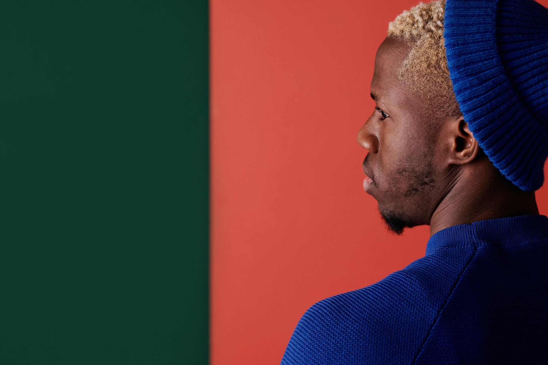 Fashion portrait of a man looking over his shoulder, wearing a blue beanie against a red and green backdrop.