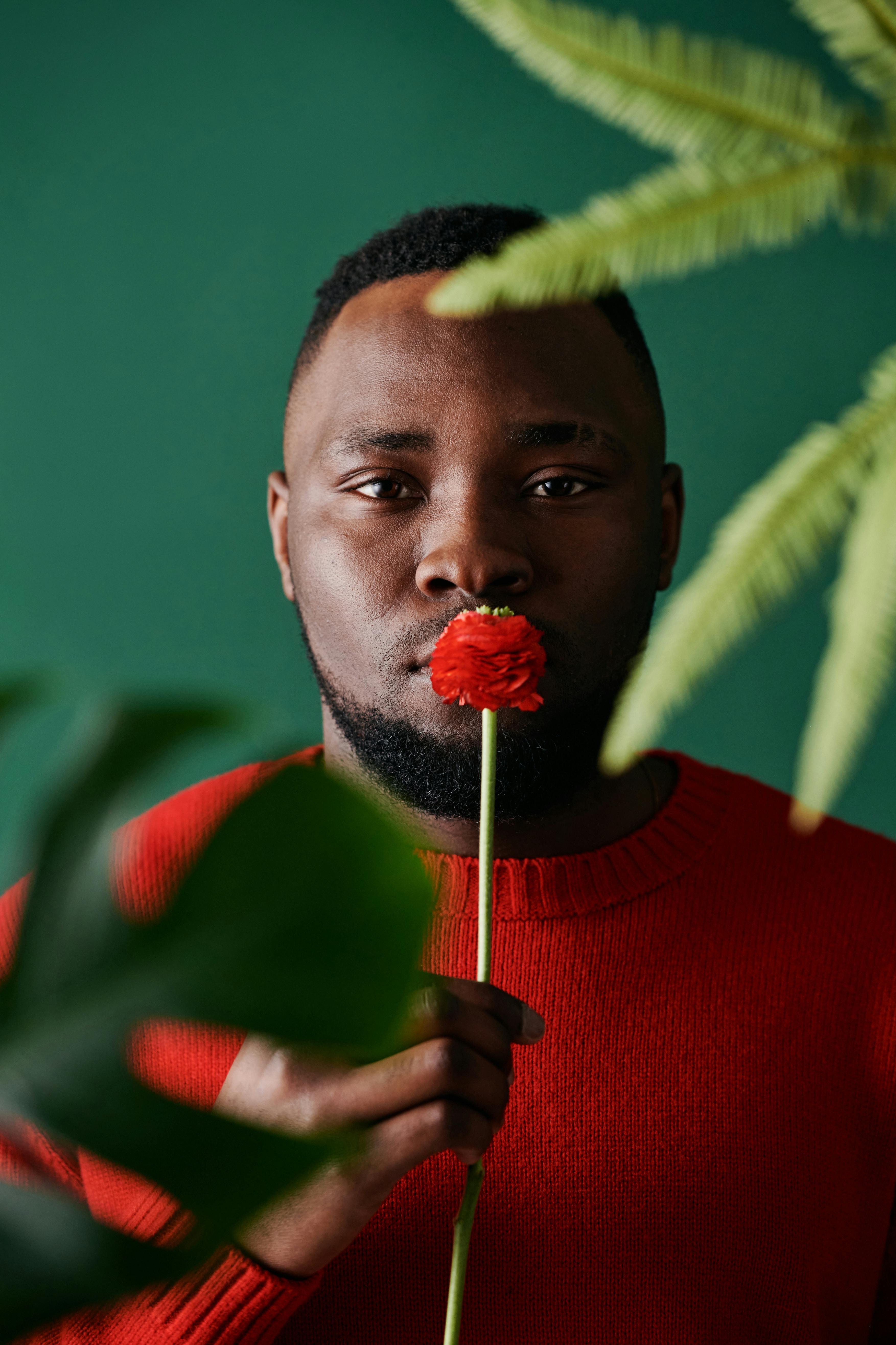African American man in red sweater holding a flower, surrounded by green leaves.