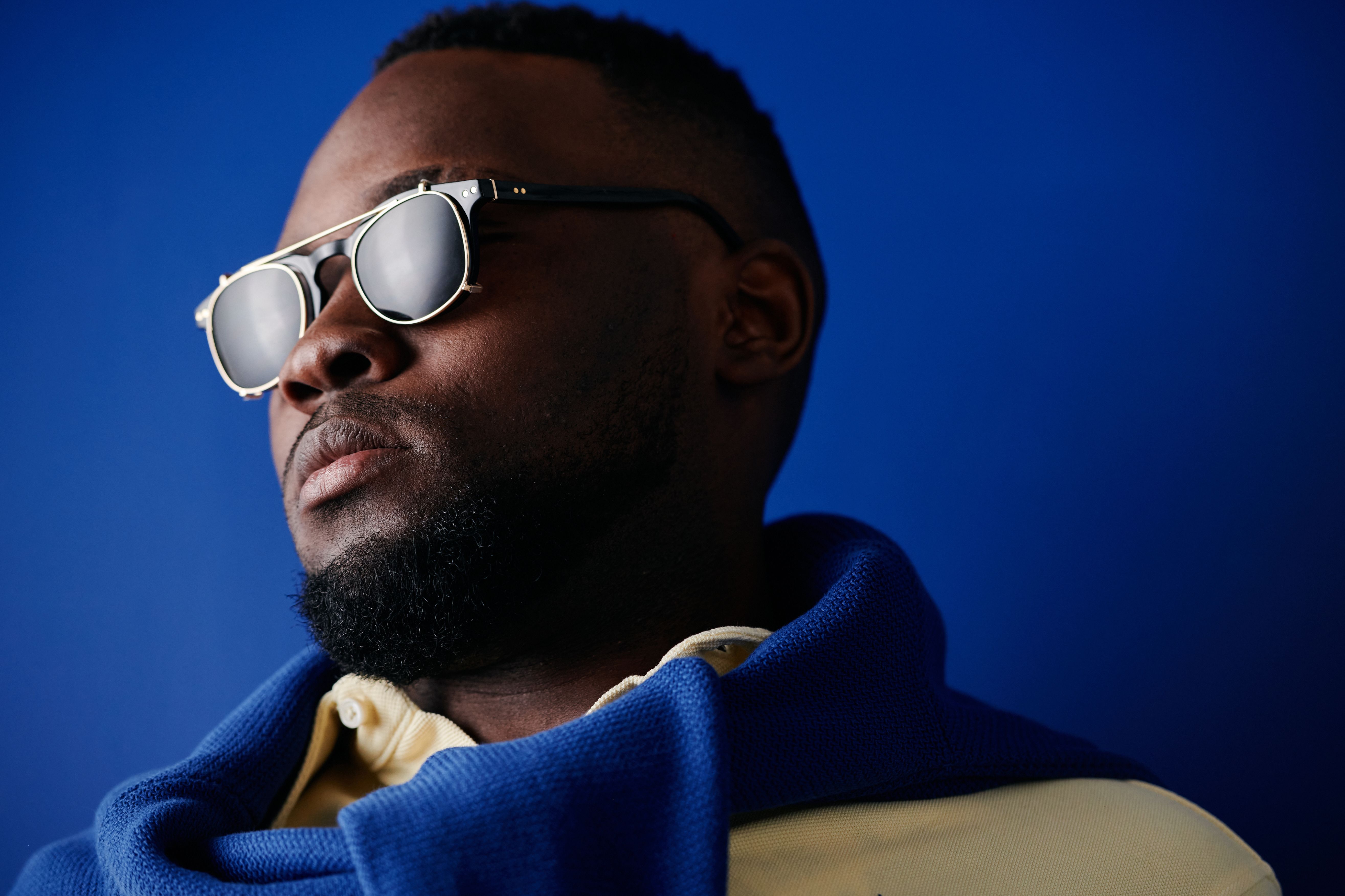 Portrait of a bearded man wearing sunglasses and a scarf against a deep blue background.