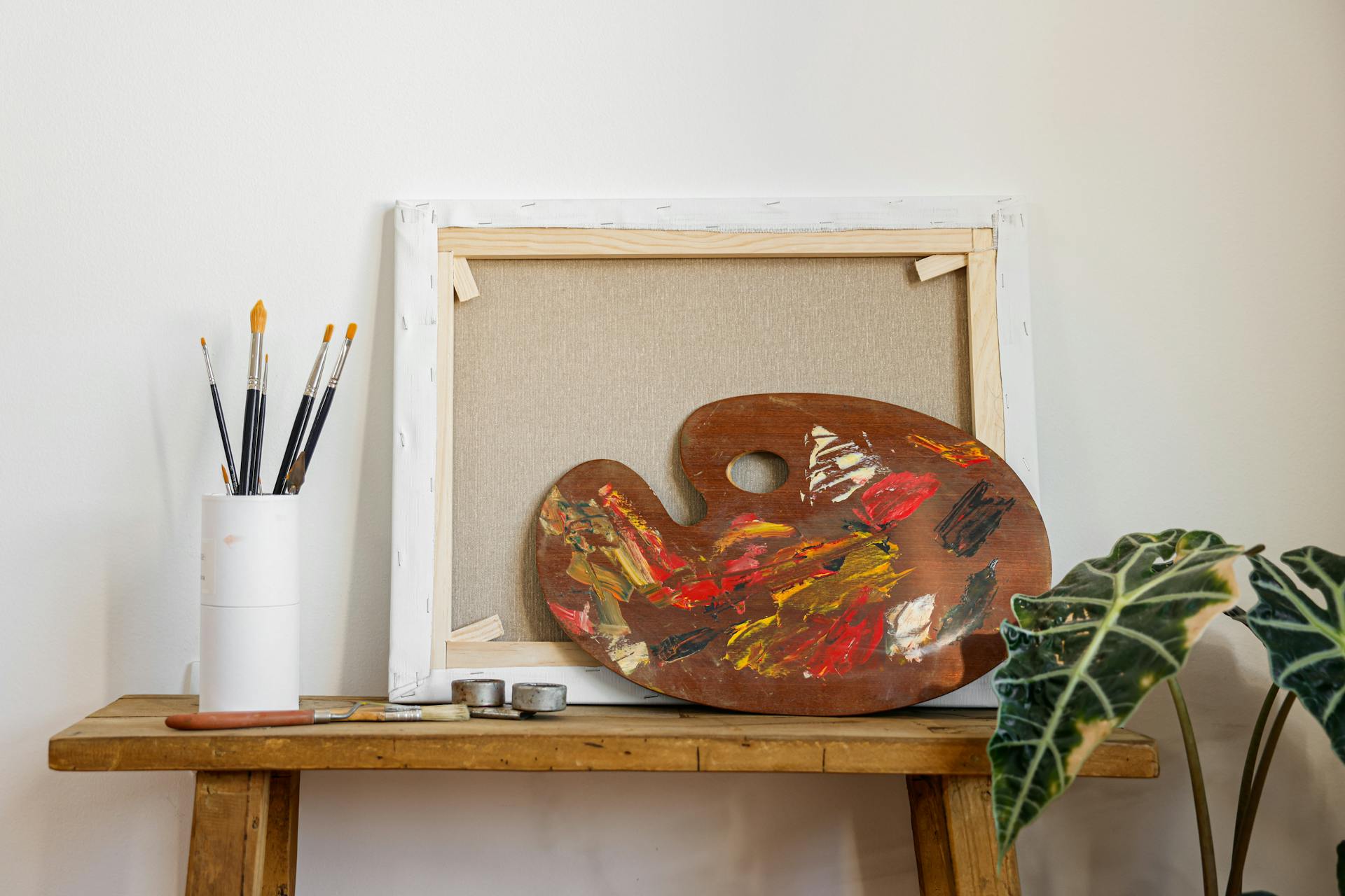 Colorful artist palette and brushes on a wooden bench beside a blank canvas and indoor plant.