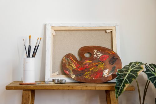 Free Colorful artist palette and brushes on a wooden bench beside a blank canvas and indoor plant. Stock Photo
