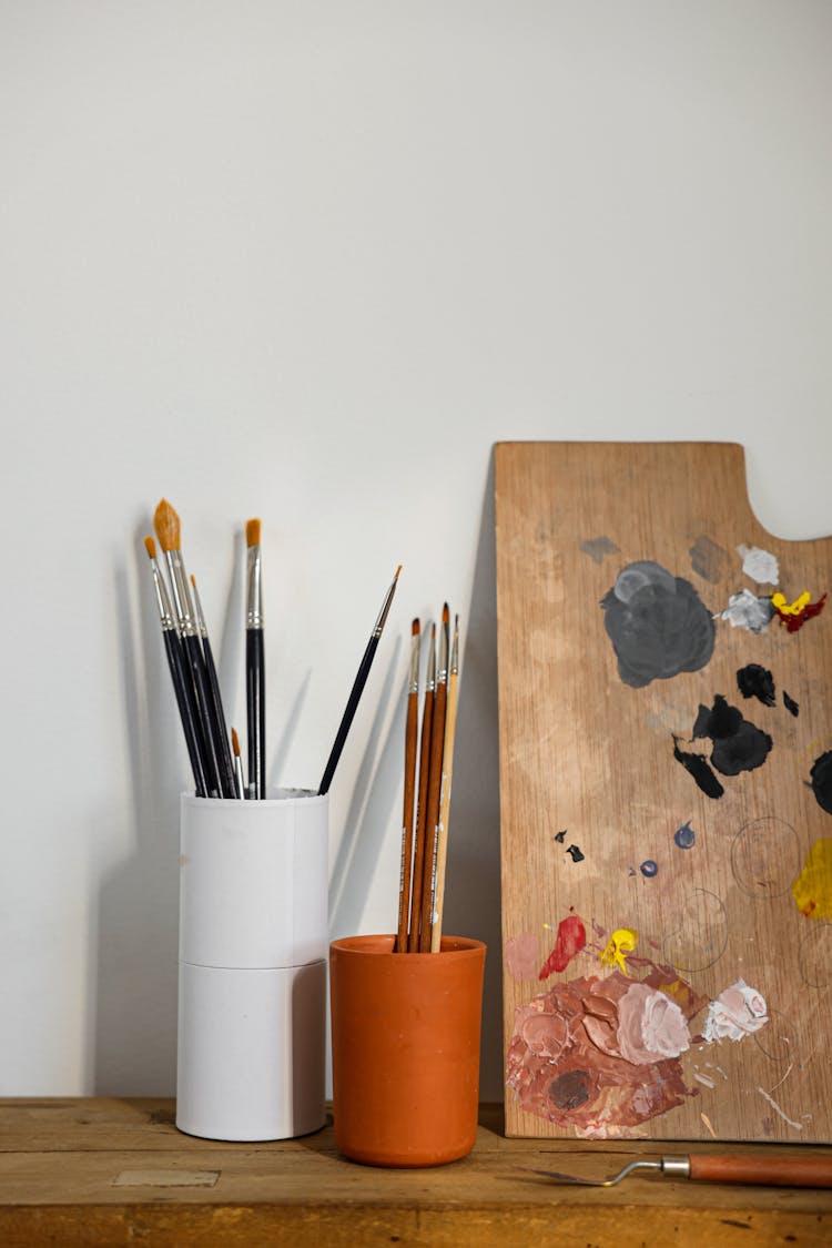 Paint Brushes In White And Brown Cups Beside A Wooden Palette