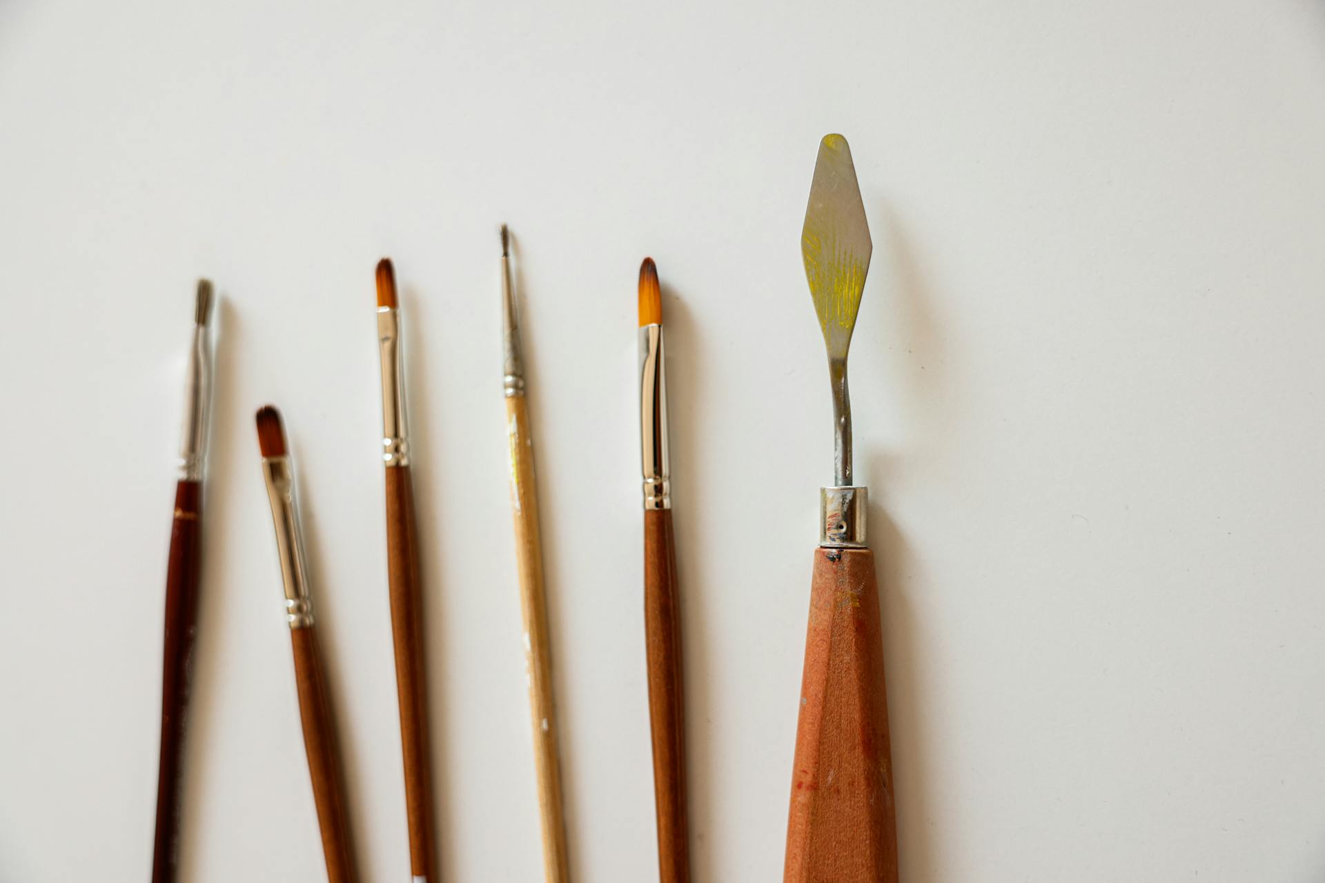 Lineup of paint brushes and a palette knife on a neutral background.