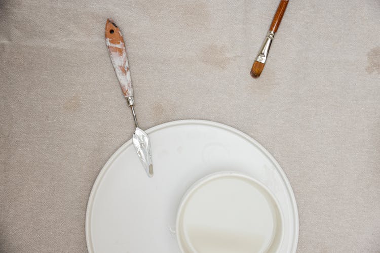 Top View Of A Paintbrush And Palette Knife With White Plates On Beige Cloth