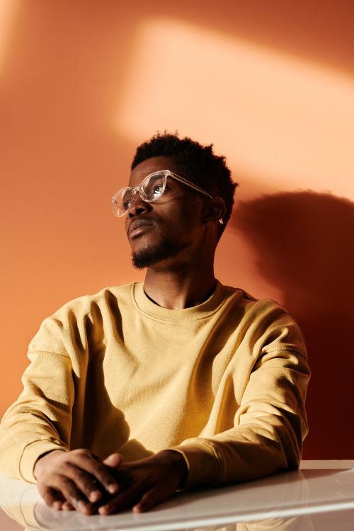 Free Fashionable portrait of a thoughtful man wearing eyeglasses and a yellow sweatshirt, sitting indoors. Stock Photo