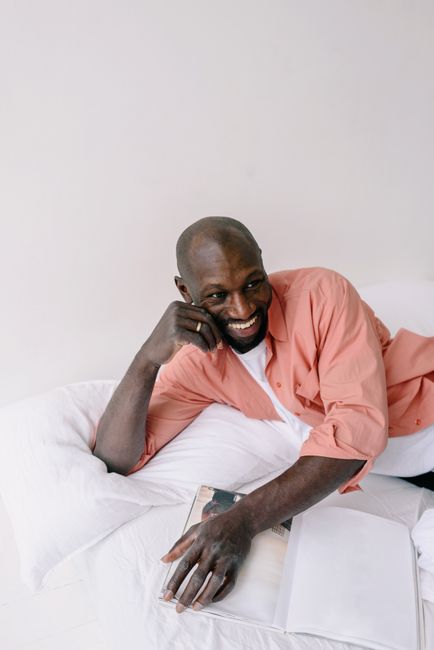 A joyful man in a pink shirt smiles while lounging on a bed with a magazine.
