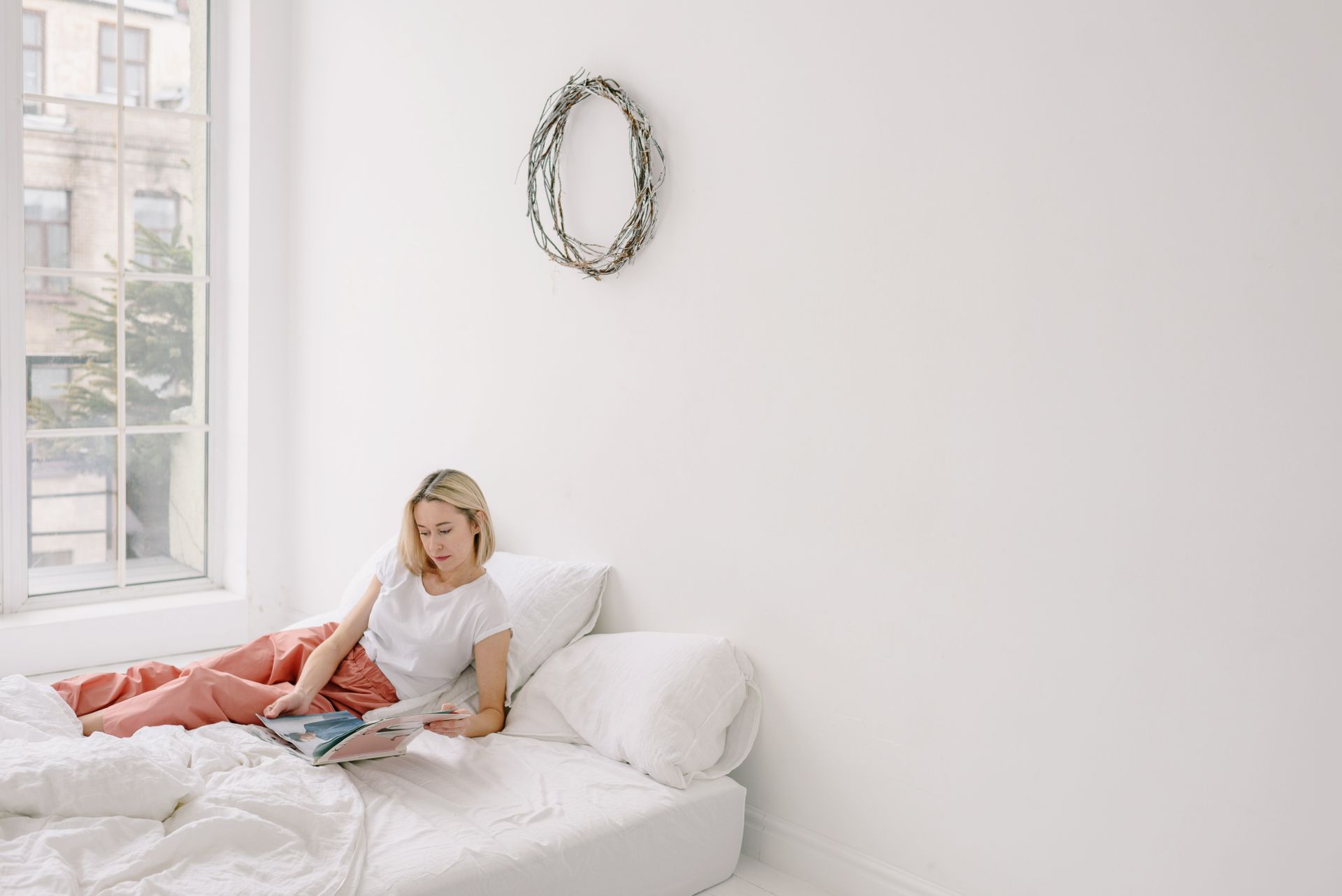 A woman enjoys a peaceful morning reading in a bright, minimalist bedroom with soft light.