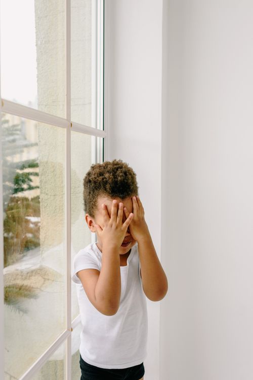 Free Cute child plays peekaboo, covering face with hands by a bright window, wearing a white t-shirt. Stock Photo