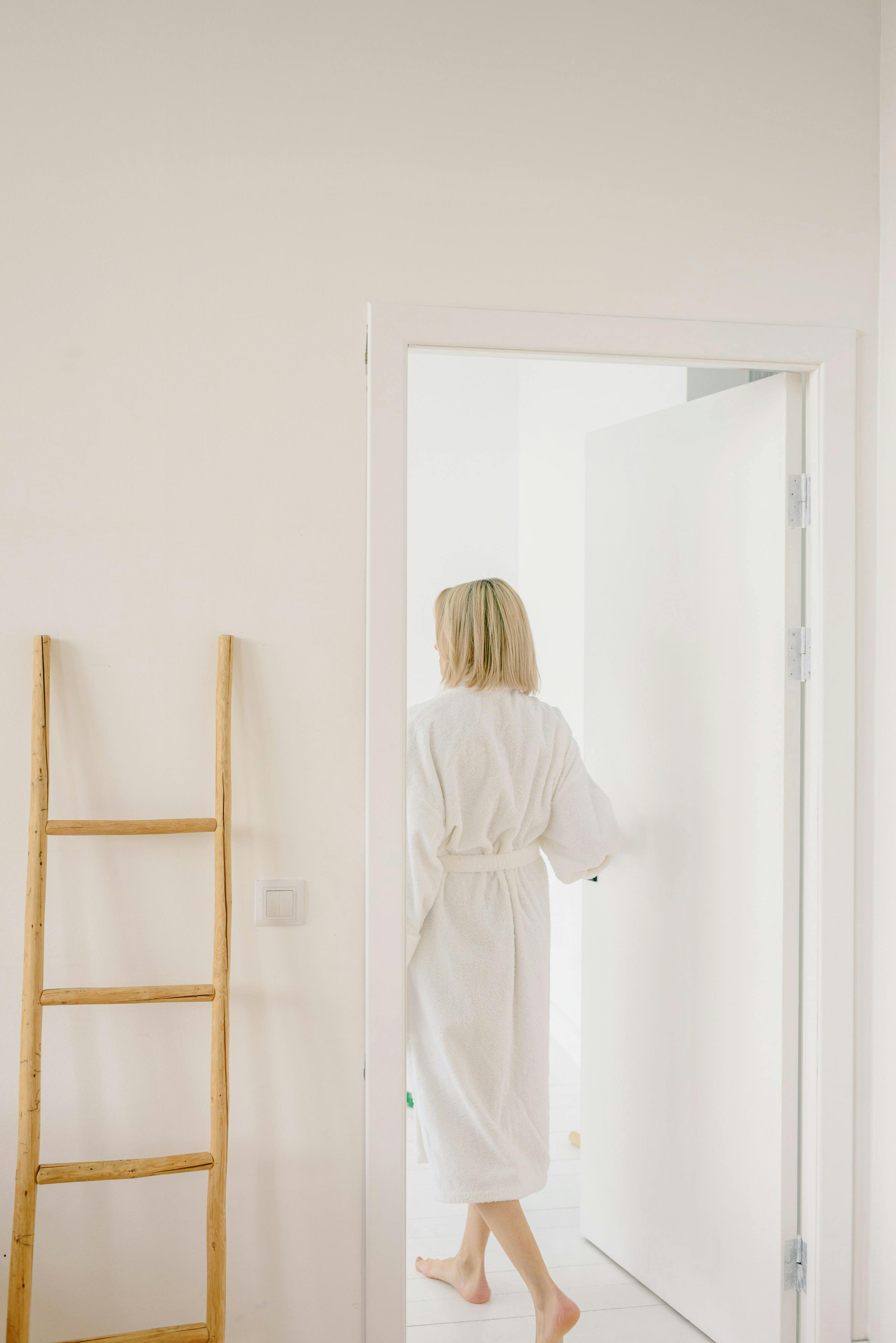 A woman in a white bathrobe enters a minimalistic, bright room softly lit by natural light.