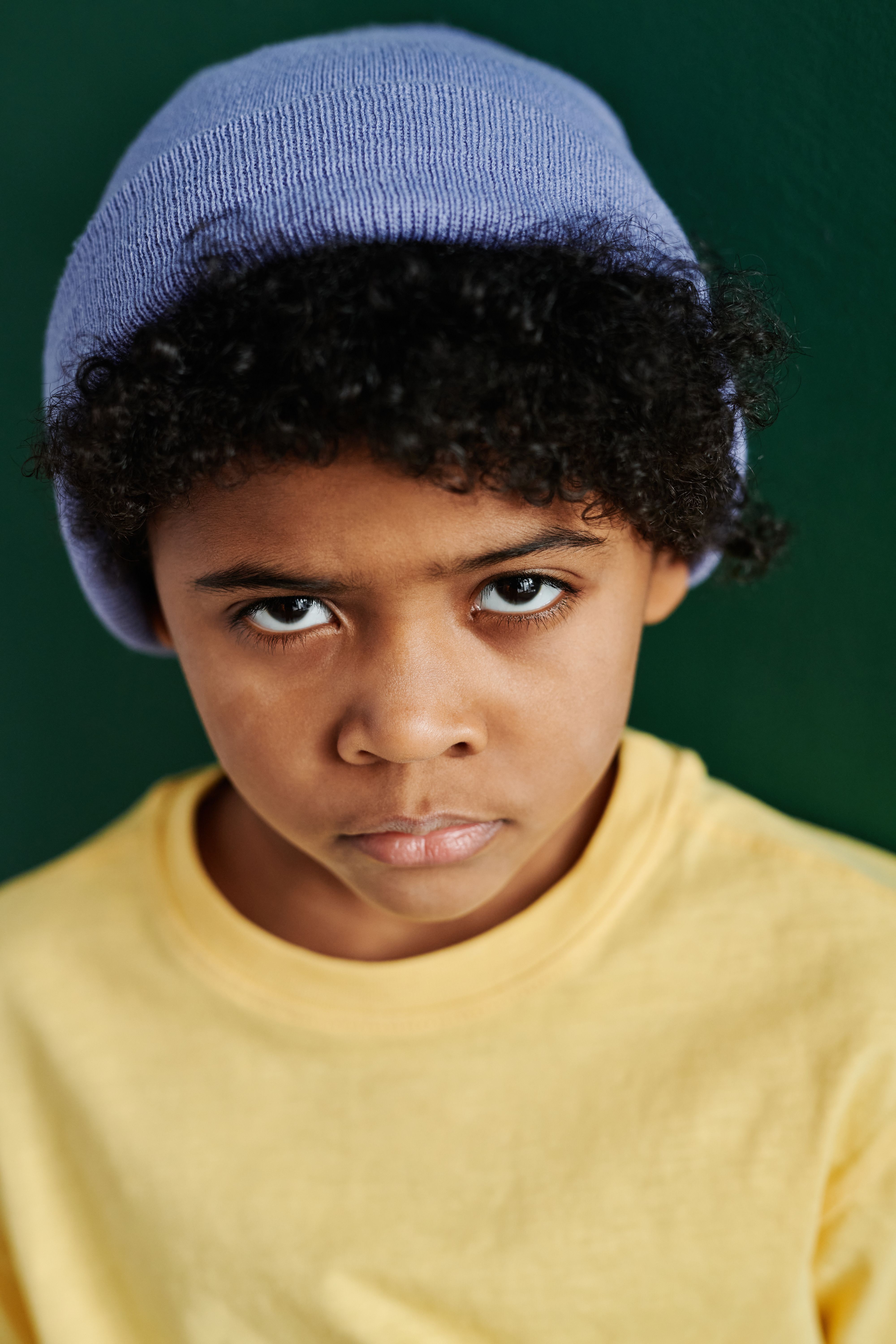 Focused portrait of a kid wearing a blue beanie and yellow shirt, against green background.