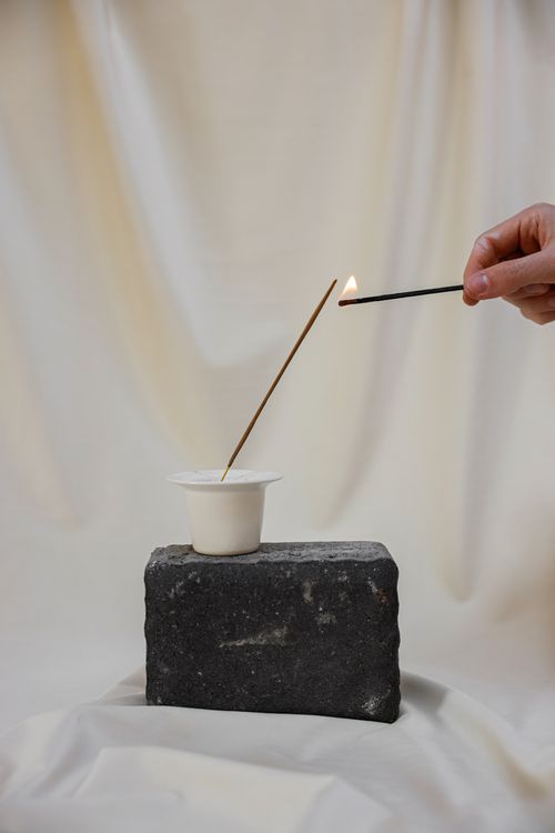 Free A hand lighting an incense stick with a match above a cup on a stone block against a neutral background. Stock Photo