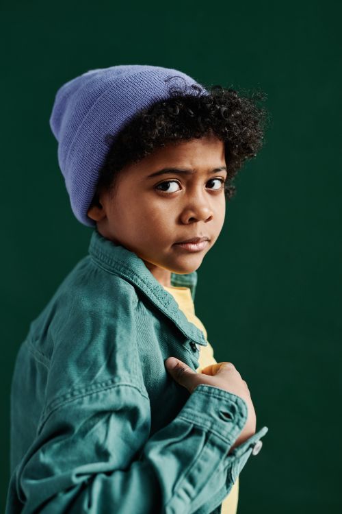 Free Portrait of a fashionable young boy wearing a blue beanie and green jacket against a dark background. Stock Photo