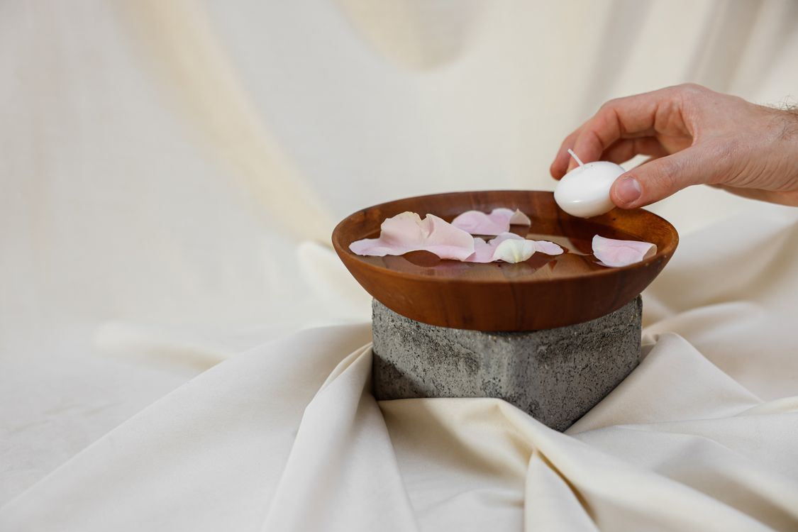 A hand placing an unlit candle in a wooden bowl of water with rose petals, a no-fire setup for a full moon release ritual