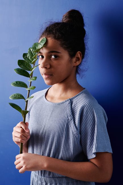 A young girl with a leaf in front of her face stands against a blue backdrop, displaying an artistic vibe.