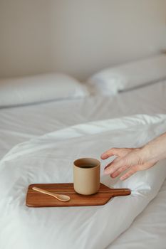 A hand reaches for a warm cup of coffee on a bed with white sheets.