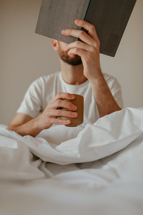 Free Man Sitting in Bed, Reading and Book and Drinking Coffee Stock Photo