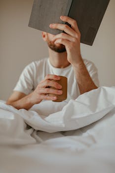 A serene morning scene of a man drinking coffee and reading in bed, creating a peaceful atmosphere.