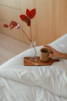 A hand holding a cup of coffee on a bed with red leaves in a vase.