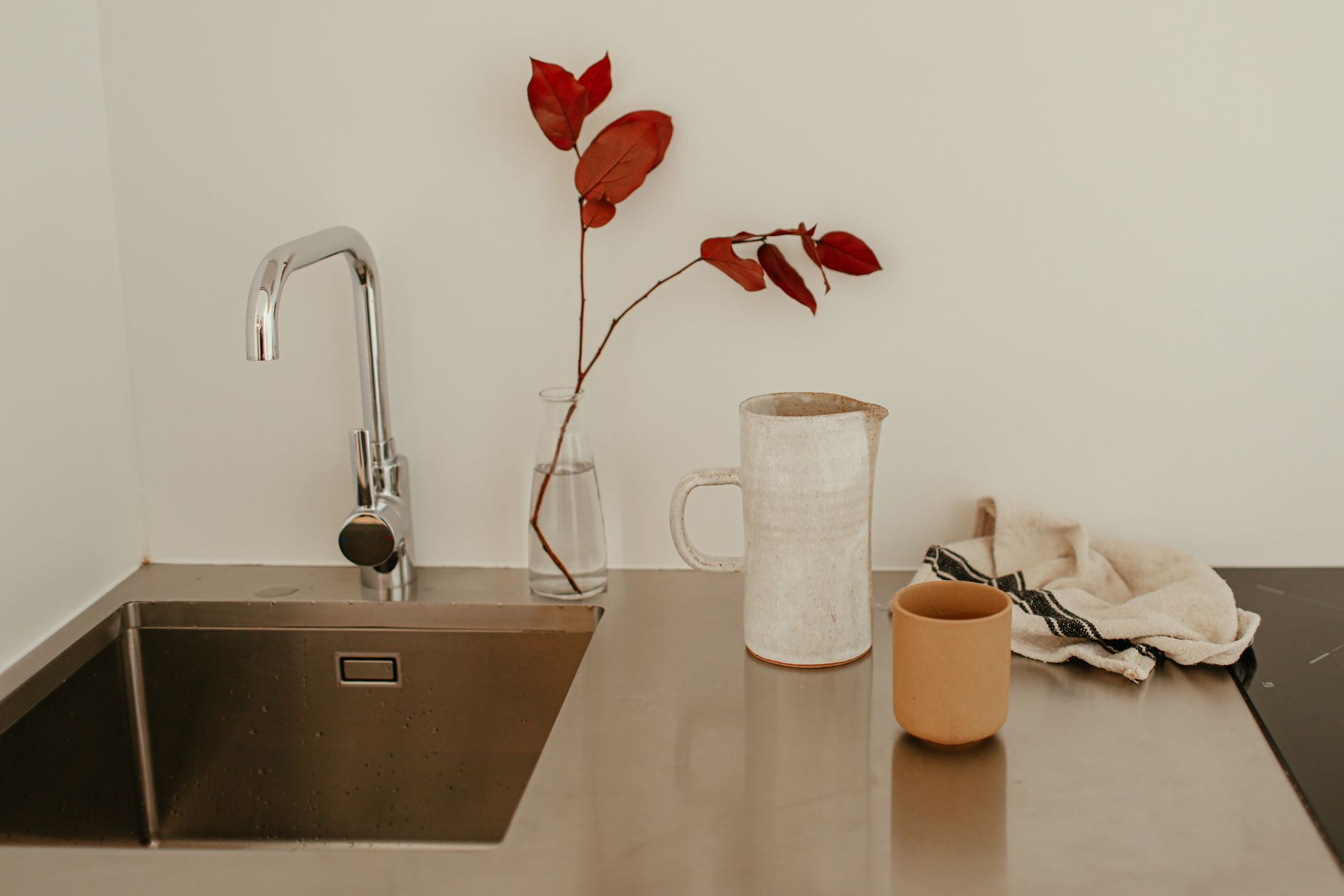 A modern minimalist kitchen sink setup with a ceramic pitcher, red leaves, and a cup on a stainless steel countertop.