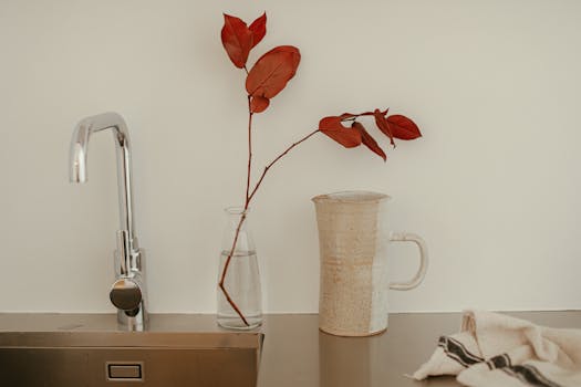 Elegant minimalist kitchen scene featuring a faucet, vase with leaves, and textured pitcher.
