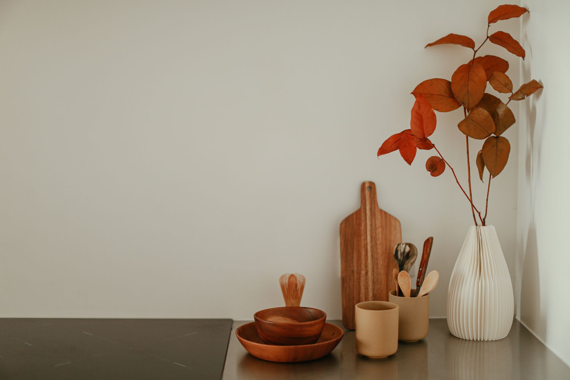 A minimalist kitchen corner with wooden bowls, a vase with autumn leaves, and cooking utensils on a countertop.