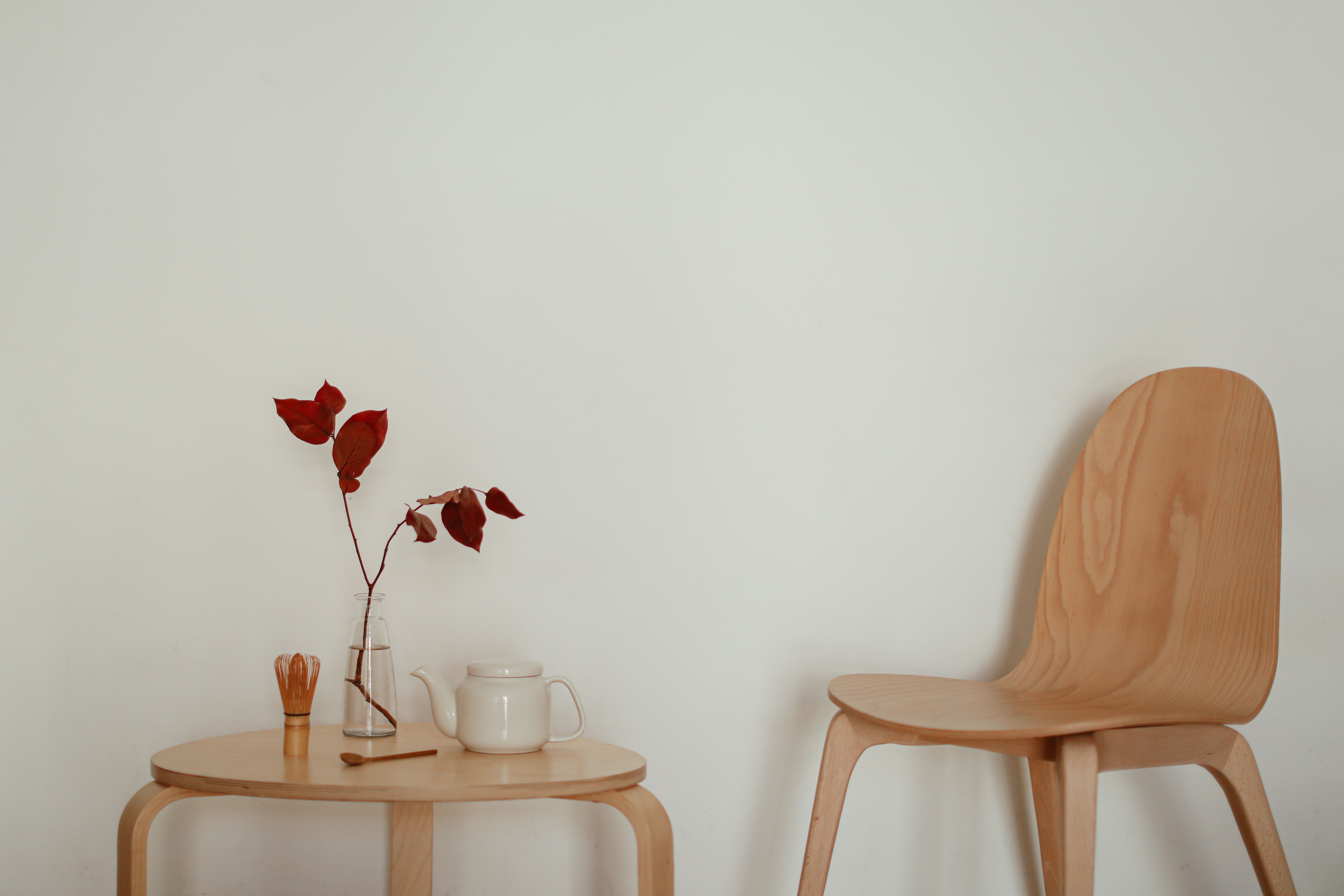 A minimalist interior featuring a table with red leaves in a vase, a teapot, and a wooden chair against a neutral wall.