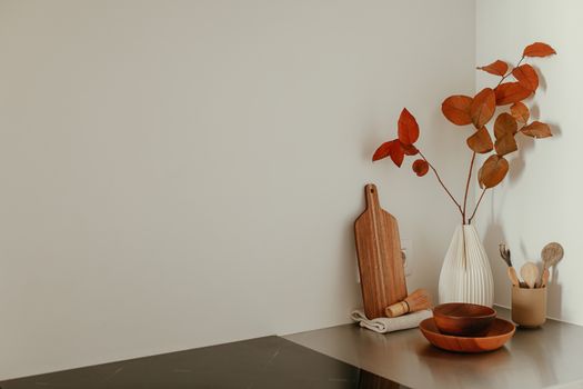 A minimalist kitchen corner featuring red leaves in a ceramic vase, wooden utensils, and a chopping board.