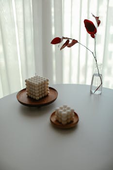 A minimalist still life featuring geometric candles on wooden stands with a glass vase and red foliage.