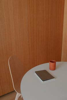 Modern minimalist workspace with a notebook and cup on a round white table against a wooden backdrop.