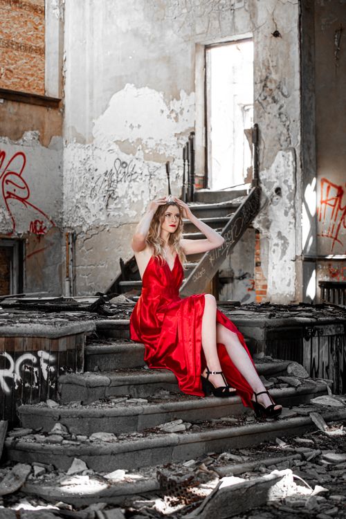 Free A woman in a red dress sits on stairs, surrounded by urban decay in an abandoned building. Stock Photo