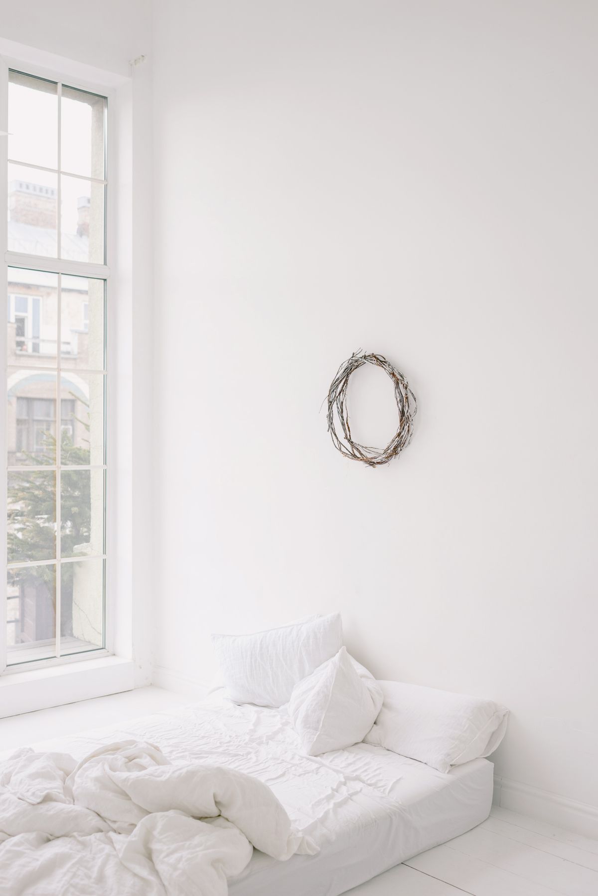 Bright minimalist bedroom featuring a white bed, large window, and wall wreath. Stock Photo.