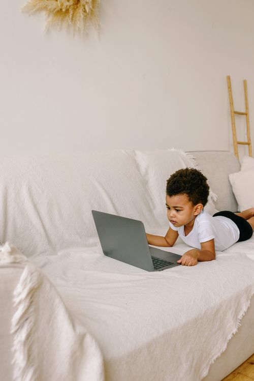 Free A child with curly hair uses a laptop while lying on a white sofa indoors. Stock Photo
