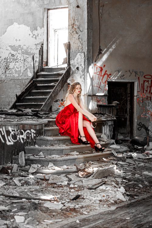 Free A young woman in a red dress sits gracefully in a derelict building, blending elegance with decay. Stock Photo