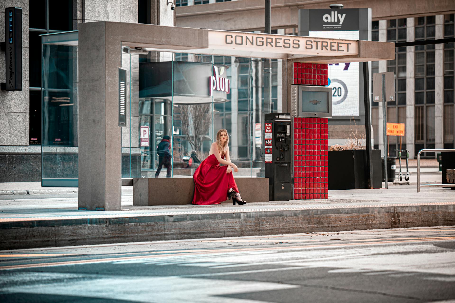 A stylish woman in a red dress sits at a Detroit bus stop on Congress Street.