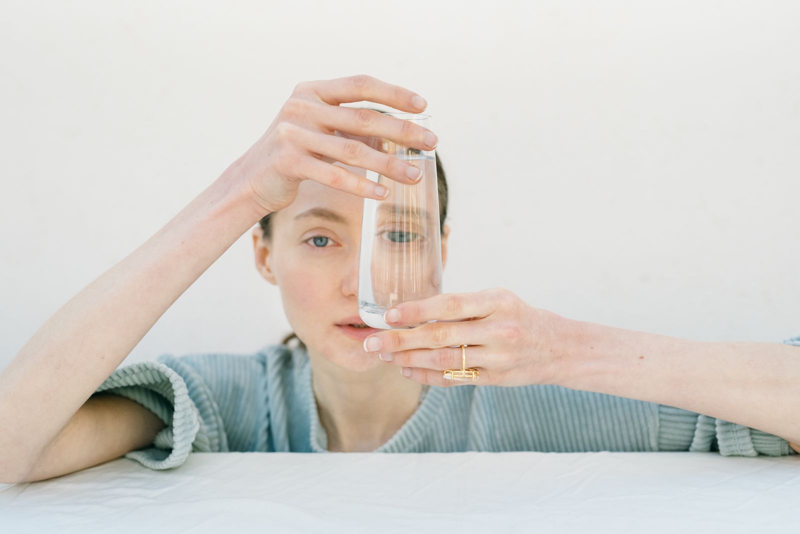 Woman with hair pulled back holds a clear glass of water in front of her face against a white wall