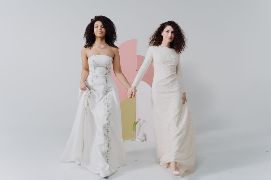 Two stylish bridesmaids dressed in elegant gowns hold hands in a bright studio setting.